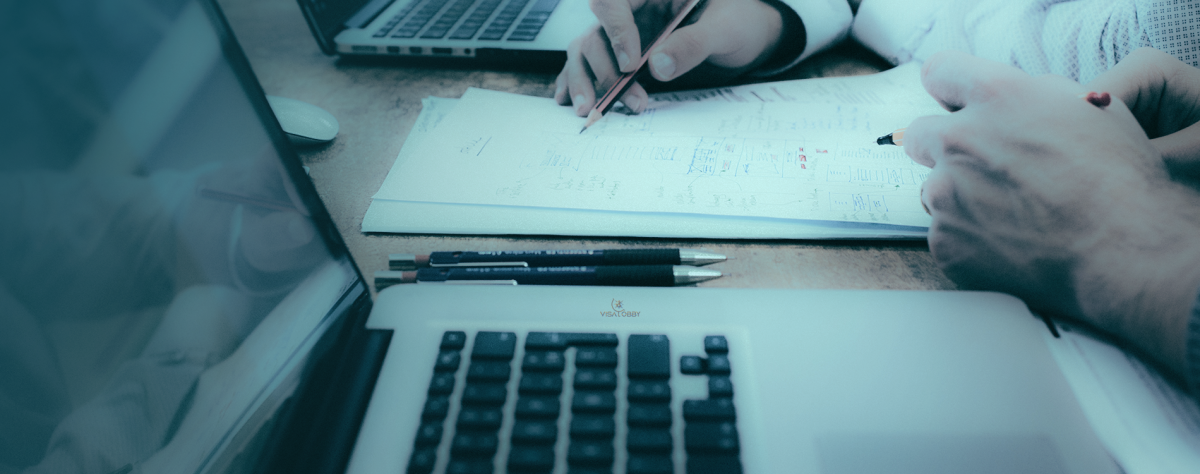 Two people are working on paper, holding the pencil in their hands. Besides, a laptop and two pens.